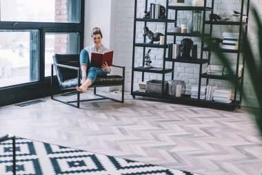 Neşeli genç bir kadın sandalyeye uzanıp gülerken evde kitap okuyor. Modern apartmanda en çok satan edebiyat kitaplarından hoşlanan pozitif hipster öğrenci.