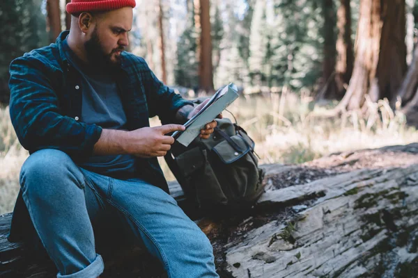 Pensive male traveler reading paper map while resting on log during trekking tour to forest in National Park, caucasian guy wanderlust spending weekend actively for exploring wild environment