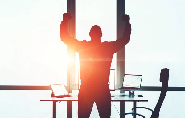 Silhouette of young businessman celebrating achievement and complete of goals raising up arms in office interior front computers desktop workplace. Male employee finish project.Motivation and progress
