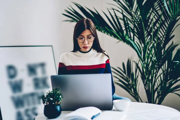 Young woman in eyewear checking email while working on freelance via modern laptop computer using internet connection at cozy apartment, hipster girl chatting online on websites for meeting people