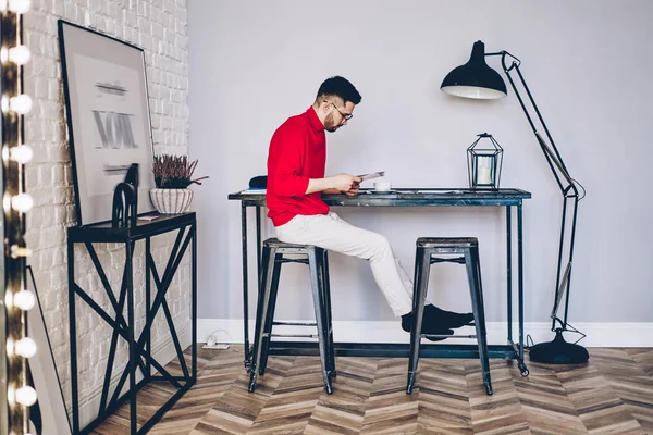 Young man reading paper documents while sitting on chair in modern apartment with stylish interior, concentrated hipster guy dressed in casual wear checking bills at home during leisure time