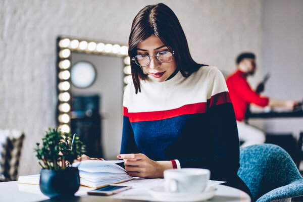 Serious pensive woman in optical eyeglasses for vision correction sitting at desktop and reading information from textbook for education during time for studying in modern flat with stylish design