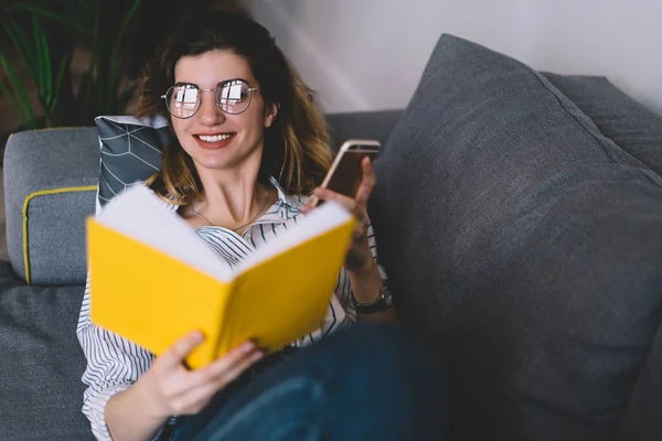 Vidám Hipster Lány Okostelefonnal Kezében Pihentető Kanapén Olvasás Kedvenc Könyv — Stock Fotó