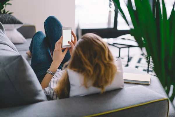 Back view of young woman lying on comfortable sofa in own room with stylish interior and chatting online with friends on modern smartphone with blank screen area for your internet content