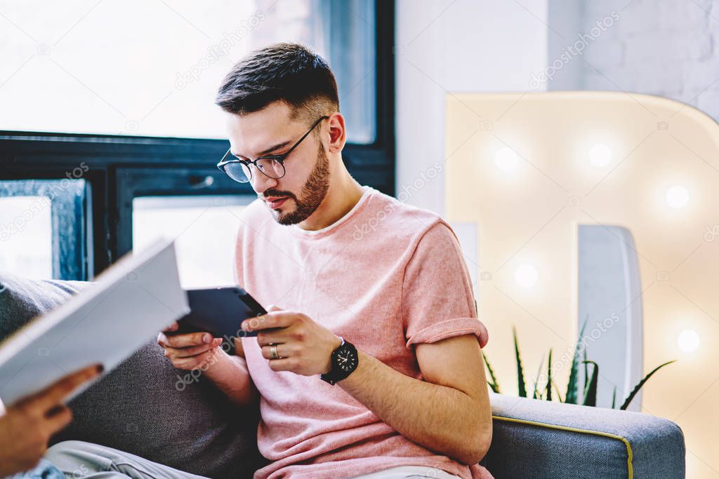 Bearded young man in eyeglasses for vision correction updating software on modern touch pad using wireless internet connection in modern apartment, male blogger downloading application at home