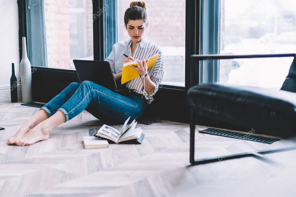 Pensive caucasian young woman holding notepad in hand and making notes for working plan sitting on floor with laptop in own apartment near window.Concentrated hipster student drawing design sketch