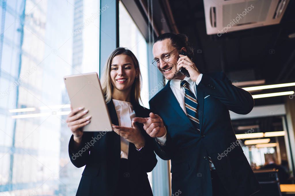 Successful cheerful male director of corporate company communicate with business partner via application on cellular phone while pointing on screen of touch pad and smiling with female colleague
