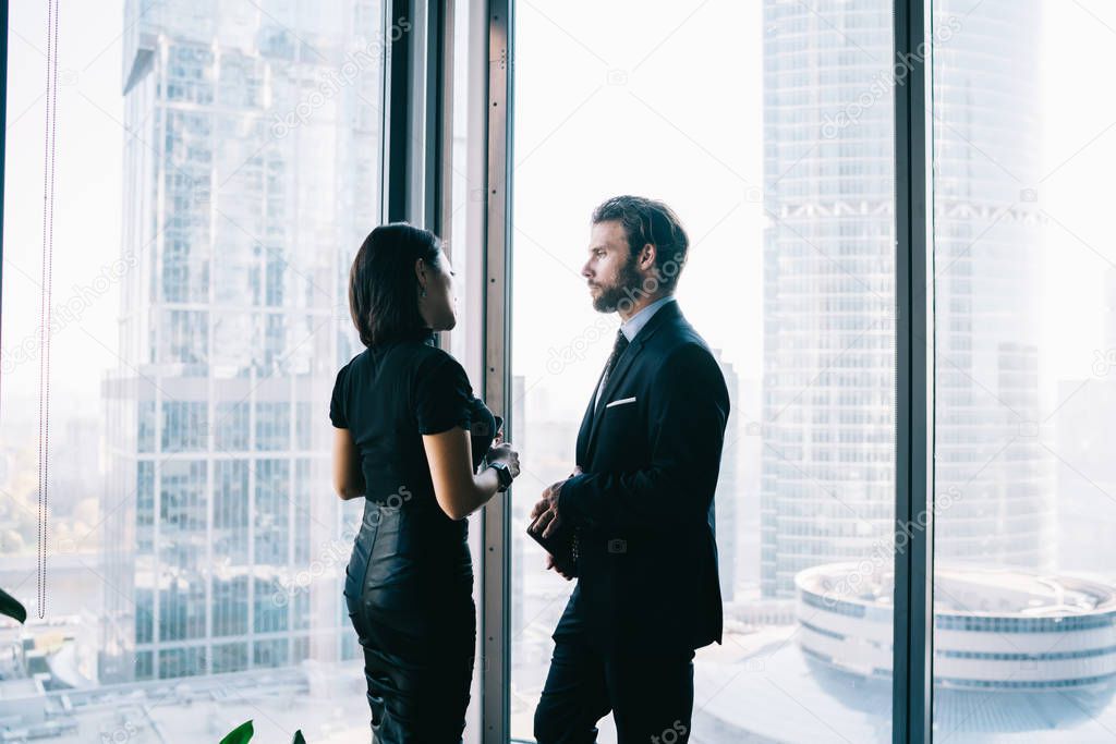 Male and female entrepreneurs dressed in elegant apparel communicating about projest ideas near window while waiting business partner for cooperation, confident proud ceo in office building