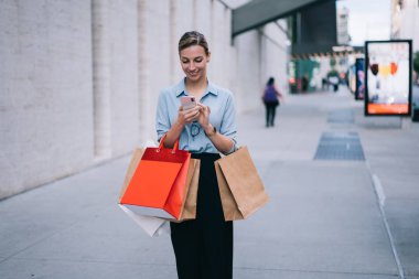 Neşeli beyaz kadın blog yazarı cep telefonuyla 4G internet bağlantısı kullanarak sosyal ağda yayın yapıyor. Alışveriş paketleriyle arkadaşlarıyla akıllı telefon aracılığıyla mesajlaşan mutlu hippi kız.