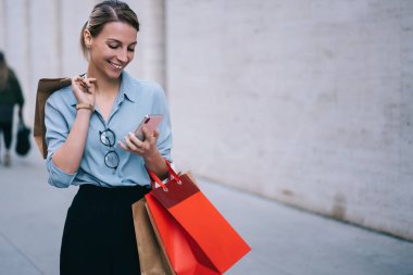 Sosyal ağda akıllı telefondan mesajlar okuyan olumlu hipster kızı şehir ortamında ayakta duruyor, satış ve indirimli mağazaları ziyaret ettikten sonra cep telefonu kullanarak gülümsüyor.