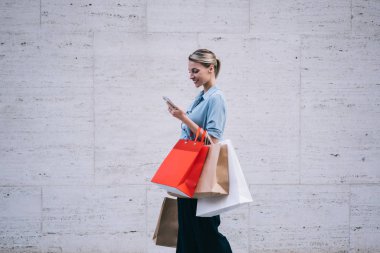 Elinde kağıt paketler olan mutlu bir kadın alışverişçi, marka adı için reklam metni için fotokopi alanının yakınında yürüyor, cep telefonunda GPS uygulaması kullanan pozitif bir kadın.
