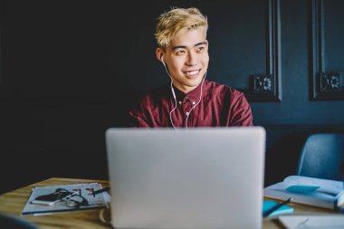 Olumlu gülümseyen Asyalı erkek öğrenci modern dizüstü bilgisayarla sohbet etmekten ve elektronik kulaklıkla güzel sesleri dinlemek için yeni müzik uygulamaları kullanmaktan keyif alıyor.