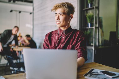 Başarılı Asyalı erkek serbest çalışan, modern dizüstü bilgisayar, eğlence ve keyif kavramı ile uzaktan çalışırken elektronik ekipmanlarla favori müzik listesini dinlerken başka yöne bakıyor.