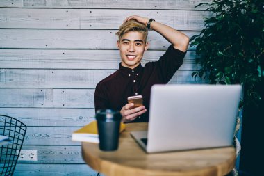 Mutlu Asyalı hippi öğrenci yüzünde sevimli bir gülümsemeyle kafeteryada dinlenirken elinde akıllı bir telefonla internete bağlı bir dizüstü bilgisayarla uzaktan çalışmaktan dolayı dinleniyor.