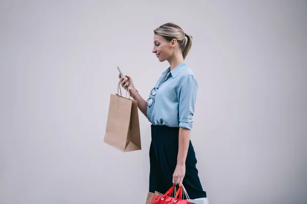 Side view of positive caucasian shopaholic with paper bags doing online shopping in web store during Black Friday sales via smartphone gadget after day in mall, woman walking near promotional wall