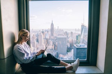 Havalı giyinmiş hippi kız otel penceresinin pervazında otururken dijital tablet kullanıyor. New York manzarası nefes kesici. Serbest çalışan kadın dijital göçebe, milenyumda çalışıyor.
