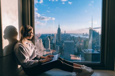Beyaz kadın kulaklıkla müzik dinlerken dizüstü bilgisayarla çalışıyor ve gökdelenlerin penceresinden New York şehrinin güzel manzarasının tadını çıkarıyor. Hippi kız turist.