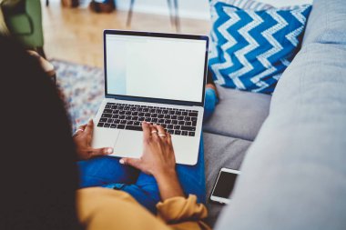 Laptop bilgisayarında yazı yazan koyu tenli bir kadının görüntüsü sahte uo-ekran görüntüsüyle evde iç mekanda zaman geçiriyor, kadın serbest çalışan uzaktan kumandalı kanepeye uzanmış, modern netbook eşiyle bağlantılı.