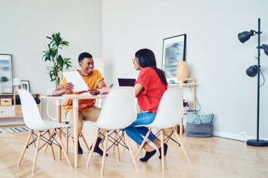 Gülümseyen koyu tenli erkek ve kadın meslektaşlar birbirleriyle işbirliği içinde konuşuyorlar, neşeli Afro-Amerikan öğrencileri modern mobilyalarla birlikte parlak bir dairede birlikte öğreniyorlar.