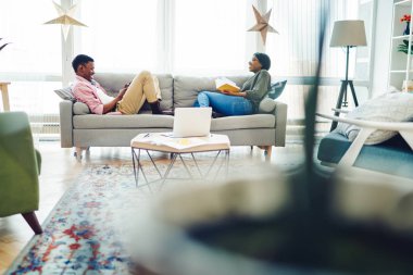 Gülümseyen Afro-Amerikan hipster çift, parlak modern iç mekanda koltukta oturuyor ve şakalaşıyor, pozitif, koyu tenli genç bir evlilik, hafta sonları rahat apartman dairesinde eğleniyor.