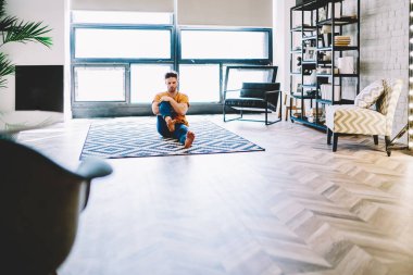Modern apartman dairesinde rahat bir evde rahat bir halının üzerinde oturan fiziksel olarak eğitilmiş genç bir adam. Hippi adam evde sağlıklı bir yaşam tarzını desteklemek için yoga egzersizleri yapıyor.