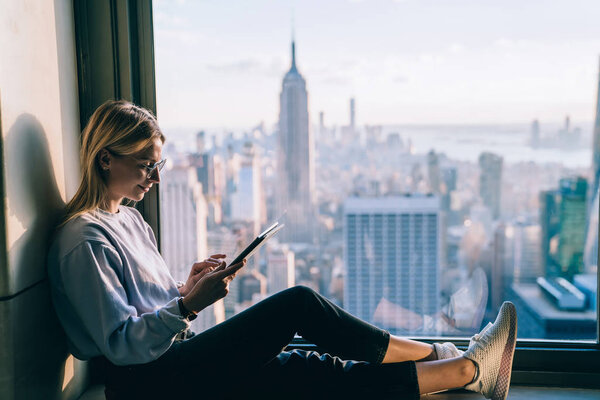 Caucasian young woman texting message on touch pad application while sitting front window with Empire State Building sight view. Female tourist enjoying roaming internet connection while travel to NYC