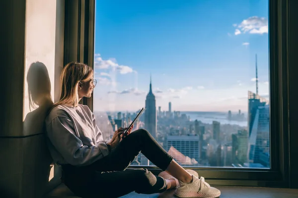 高層ビルの高層階にあるホテルの部屋に座っている間 ニューヨークの美しい景色を楽しむ笑顔のヒップスターの女の子 休暇中の通信のためのタッチパッドを使用して幸せな女性観光客 — ストック写真