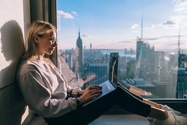 Caucasian female digital nomad keyboarding article for travel blog while working on laptop computer in modern coworking office with panoramic views of New York downtown. Woman tourist at window sill