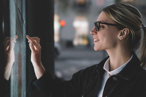 Smiling female standing at big display with advanced digital technology. Young woman touching with finger sensitive screen of interactive kiosk for find information while standing on street in evening