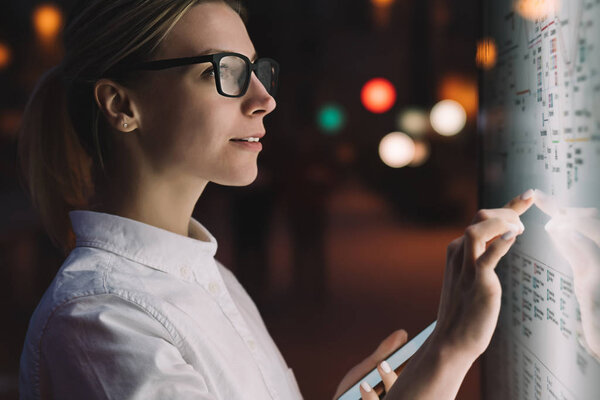 Digital monitor with public transport subway map. Female standing at big display with smartphone in hand. Young woman touching with finger screen while using train schedule application on mobile phone