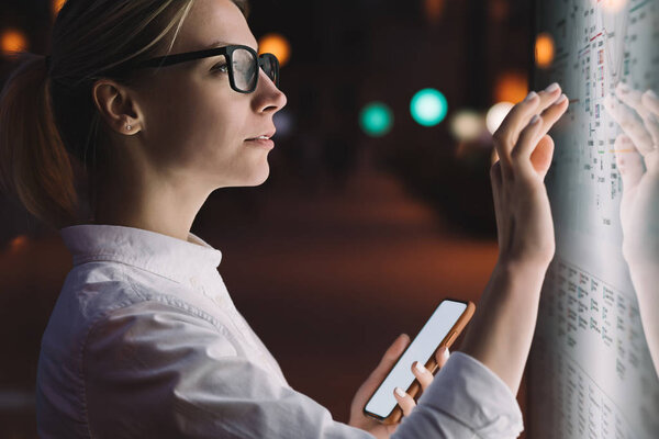 Interactive kiosk with public transport subway map.Female standing at big display with smartphone in hand.Young woman touching with finger screen while using train schedule application on mobile phone