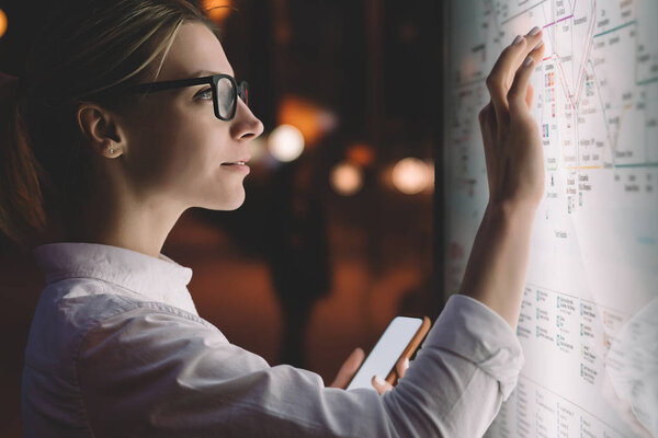 Interactive kiosk with public transport subway map.Female standing at big display with smartphone in hand.Young woman touching with finger screen while using train schedule application on mobile phone