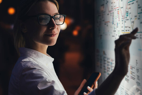 Interactive kiosk with public transport subway map.Female standing at big display with smartphone in hand.Young woman touching with finger screen while using train schedule application on mobile phone