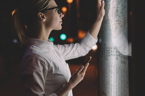 Digital monitor with public transport subway map. Female standing at big display with smartphone in hand. Young woman touching with finger screen while using train schedule application on mobile phone