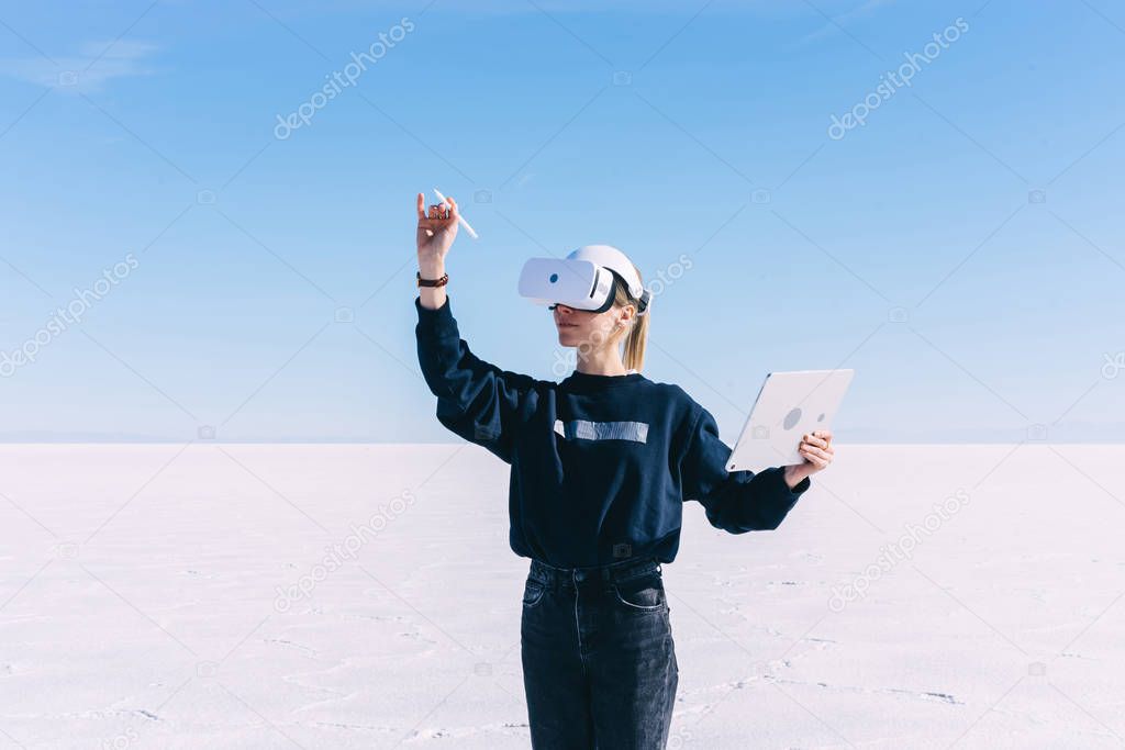 Young women extends hand with digital pencil for interactive touch over air while standing in spacious clear outdoors space wearing virtual reality headset. Hipster girl designing architectural space