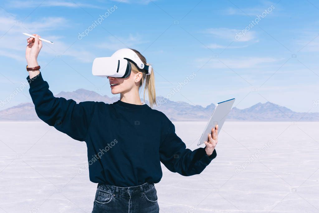 Smiling caucasian female extends hand with digital pencil for interactive touch over air while standing in futuristic outdoors space. Woman Work with 3D City Model in VR headset, innovative tech app