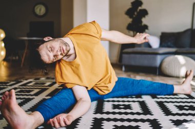 Gözleri kapalı genç bir adam halının üzerinde oturmuş, esneme egzersizleri yapıyor ve evde esneklik kazanıyor. Yoganın motive olmuş erkek aşığı, apartman dairesinde sabah antrenmanı yapıyor.