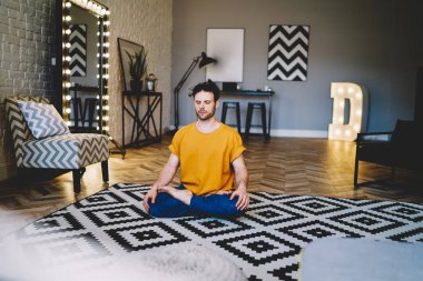 Yoğunlaşmış, gözleri kapalı, uyumun ve vücut şeklinin tadını çıkaran genç bir adam. Sabah meditasyonu sırasında Lotus pozisyonunda otururken modern apartmanın iç kısımlarında dinleniyor.