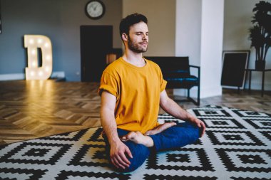 Meditasyon sırasında gülümseyen genç bir adam modern apartmanda rahat bir halının üzerinde poz veriyor. 20 yaşında, yoga yapan sakin bir hippi.