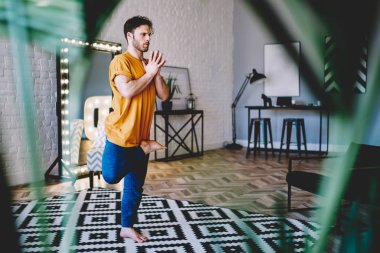 Sakin genç adam tek ayak üstünde duruyor ve namaste 'de el ele tutuşuyor. Evde sabah antrenmanı sırasında uyum ve rahatlığın tadını çıkarıyor. Yoga egzersizleri sırasında asana pozu veren konsantre hipster çocuk.