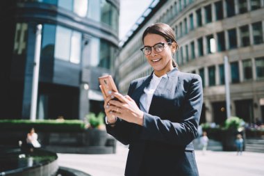 Zarif bir takım elbise giymiş, şık gözlüklü, gülümseyen, gözlüklü, neşeli kadın avukatın yarım boy portresi finans bölgesinde güneşli bir günün tadını çıkarıyor.