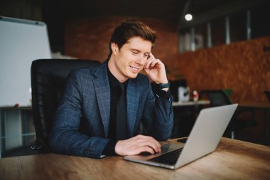 Kafkasyalı kendine güvenen erkek girişimci internet sayfasından gelen haberlerden memnun, netbook 'ta gezen gülümseyen iş adamı, netbook' taki uygulamayı kullanarak ofisindeki masaüstünde oturan muhasebe kayıtlarını kontrol ediyor.