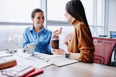 Kafkasyalı neşeli kadın meslektaşlar kahve molası sırasında arkadaşça sohbet ediyorlar. Yaratıcı projeyi birlikte gerçekleştirmekten memnun olan gülümseyen kadın çalışanlar başarılı çalışmaları tartışıyorlar.