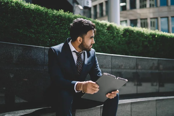 Contemplative spanish male financial expert dressed in formal suit sitting outdoors with resume in folder feeling pondering and nervous before interview meeting with corporate manager of company