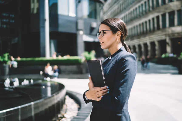 Side view of intelligent female boss looking away and thinking about ideas for productive strategy, caucasian mature entrepreneur in eyewear pondering on urban setting standing on publicity area