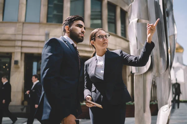 Serious male and female bankers dressed in stylish formal suits discussing architecture on publicity area, caucasian businesswoman pointing somewhere for talking with elegant spanish colleague