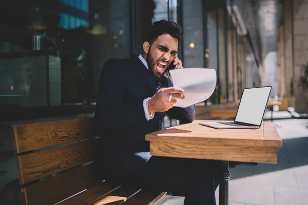 Angry turkish man in formal wear feeling rage while checking data from accounting report calling to employee and shouting for bad work while sitting outdoors with netbook with blank screen area