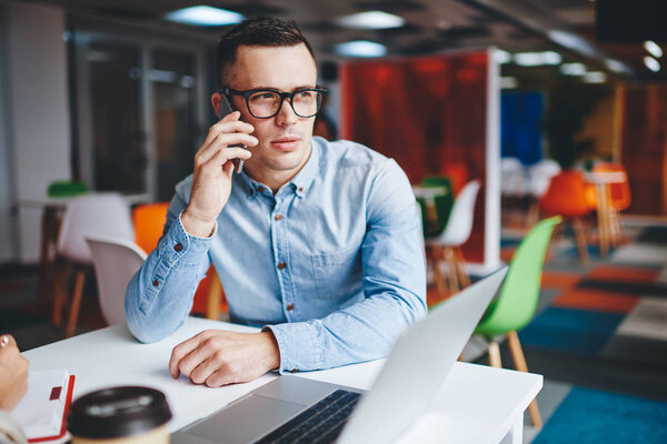 Pensive young man in eyeglasses calling on smartphone and talking with operator sitting at table with computer in office.Smart student communicating on cellular during working process at netbook
