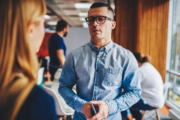 Serious young man in eyeglasses standing in office and communicating with colleague.Confident hipster student dressed in denim shirt holding smartphone and talking with friend during break in office