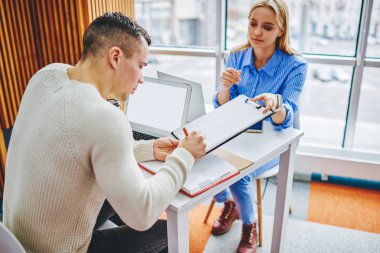 Genç bir kadın, ofis içi çalışma sırasında iş arkadaşı için belge imzalıyor. İcra Kurulu Başkanı, masaüstü bilgisayarlarıyla birlikte oturup bilgi toplamaktan gurur duyuyor.